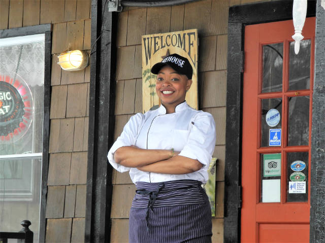 Chef Danielle Harrison, standing on the porch of the Lake House, wearing her chef coat and a Lake House baseball cap